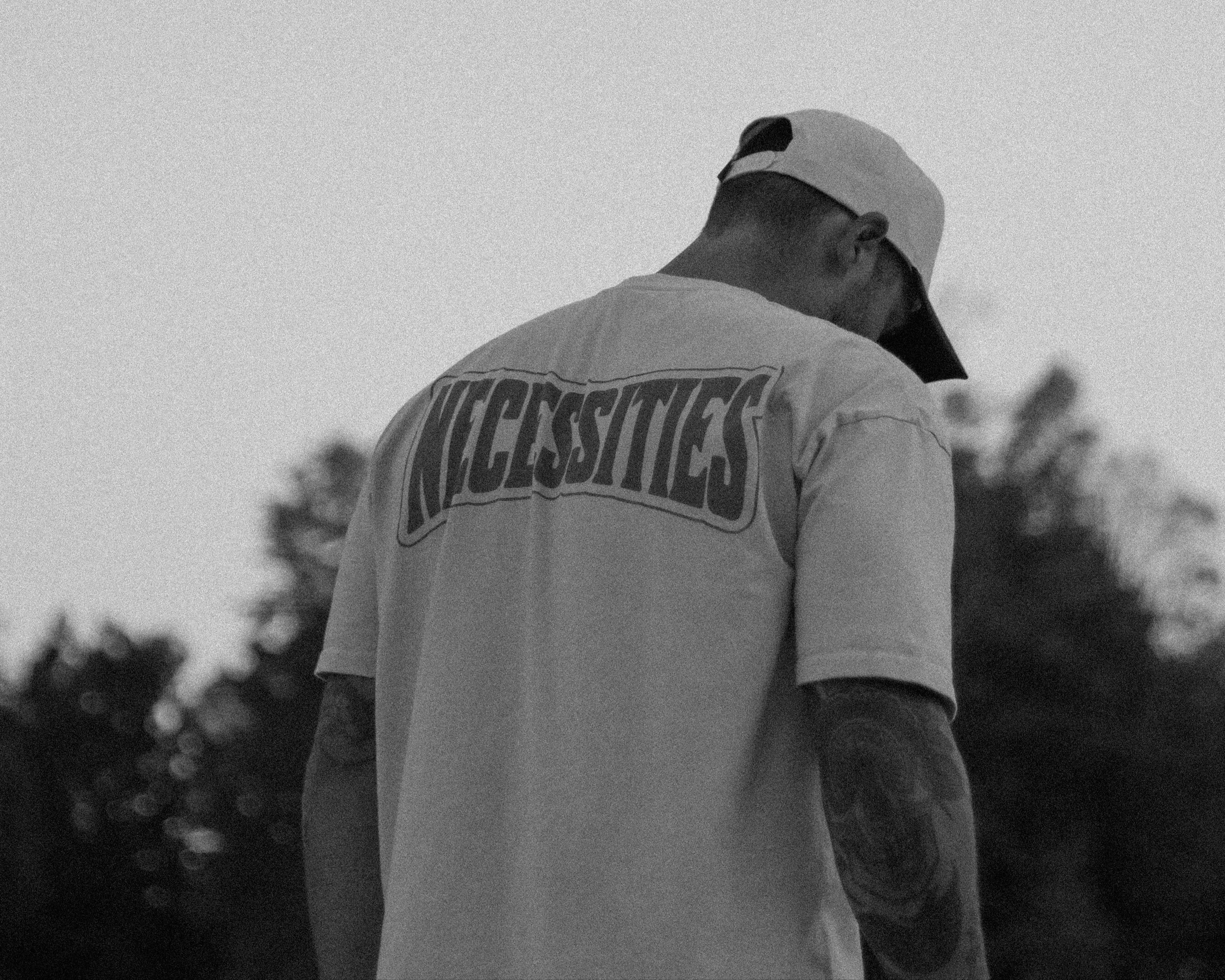 Person wearing a cap and t-shirt with 'NECESSITIES' text, standing against a sky background